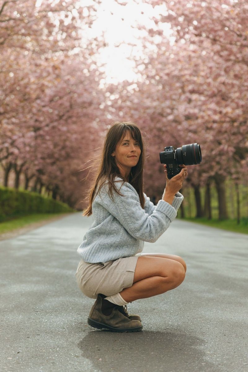 A photographer crouches on a road holding a Canon mirrorless camera, with blooming cherry trees forming a softly blurred background.