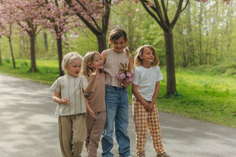 Four children walk together along a paved path lined with blossoming cherry trees, surrounded by fresh green foliage in soft natural light.