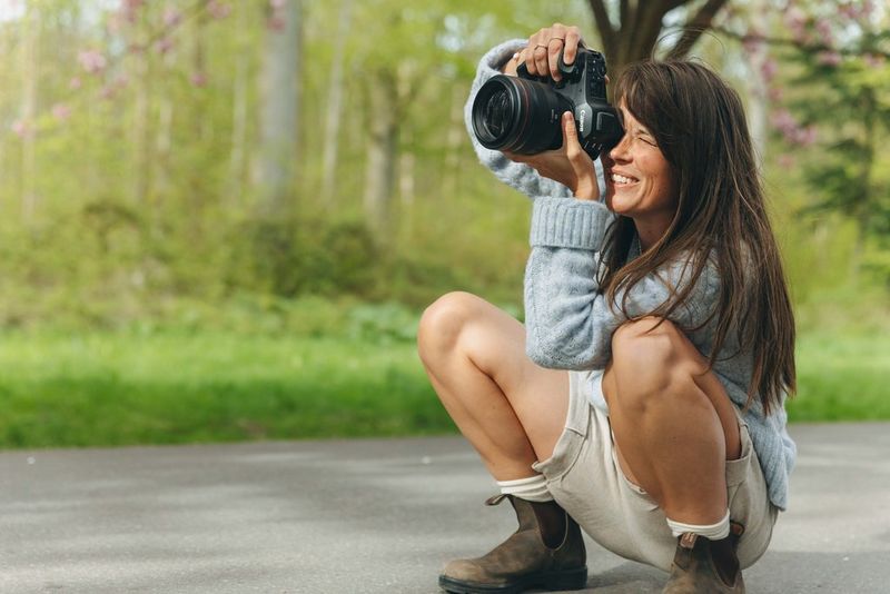 Photographer Sofie Hammer crouches on a paved path in an outdoor spring setting, holding a Canon EOS R5 Mark II up to eye level to capture a portrait. Soft natural light, fresh greenery, and trees with early spring foliage surround the scene, emphasizing a seasonal and natural approach to portrait photography.