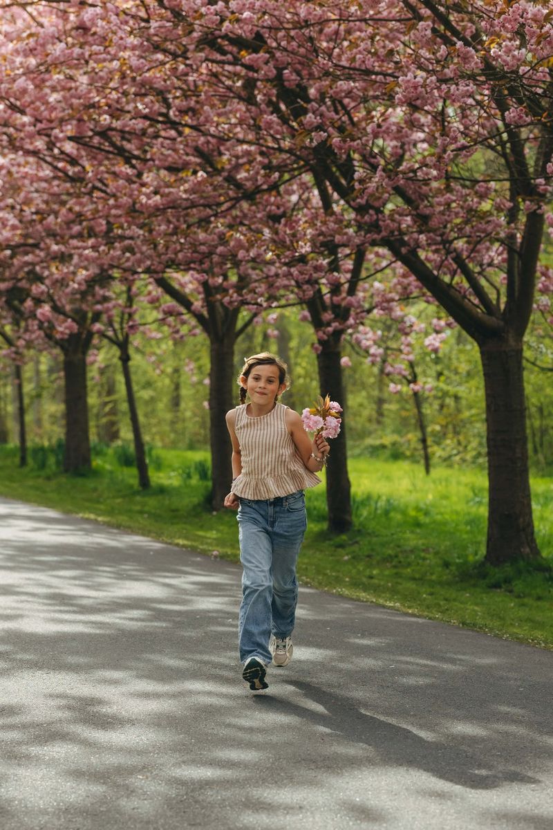 A child walks along a paved path holding cherry blossoms, with flowering trees arching overhead and green park surroundings softly blurred in the background.
