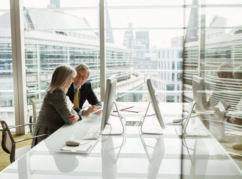 A senior businessman and woman discuss notes while sat next to a white PC and mouse on a glossy white desk.