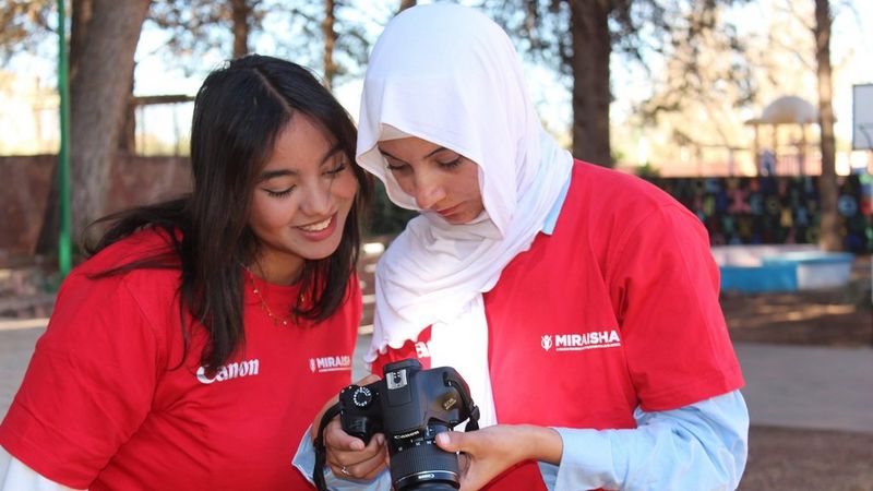 Two young women in red t-shirts with white Canon and Miraisha logos lean in together to look at the display screen of a black Canon DSLR camera.