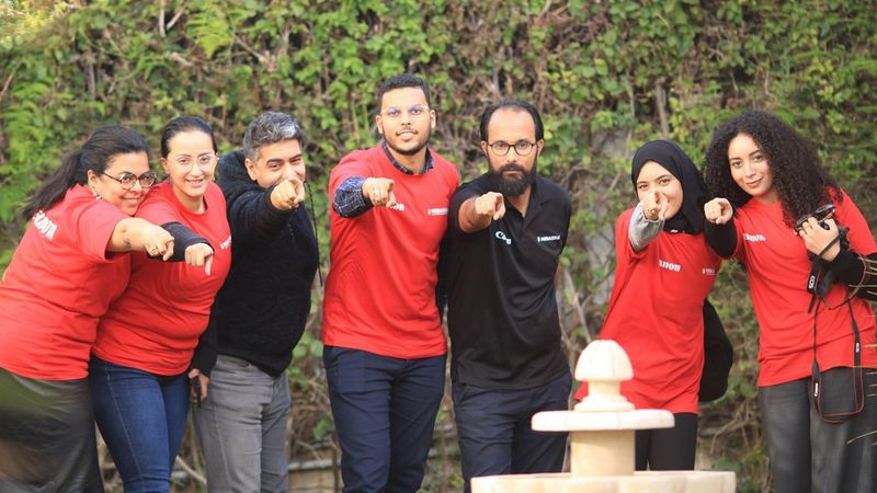 A group of seven people in red and black Canon/Miraisha t-shirts stand in a line outdoors, smiling and pointing directly at the camera.