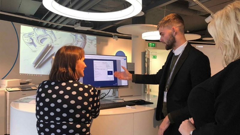 On the right, a bearded man wearing a suit gestures towards a computer screen. To his right we can see the back of a woman, who has her head facing towards him. She is wearing a black top with white polka dots. Beyond them both is a screen showing an illustration of a camera and above them is a circular light. To the far right of the shot is a partial figure of a blonde woman.