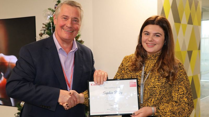 A smiling man and woman stand facing the camera, shaking hands. They are holding a framed certificate to the camera with their spare hands. The man is wearing a dark blue suit, and shirt with no tie. The woman is wearing a high-necked yellow and black blouse. They both wear lanyards around their necks.