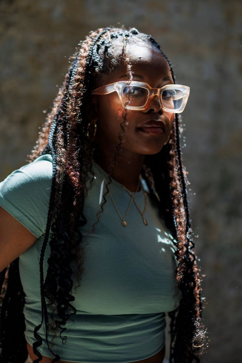 A portrait of a young woman with long braided dreadlocks taken on a Canon EOS R6 Mark II by available light, her glasses appearing to glow as they catch what little light there is.