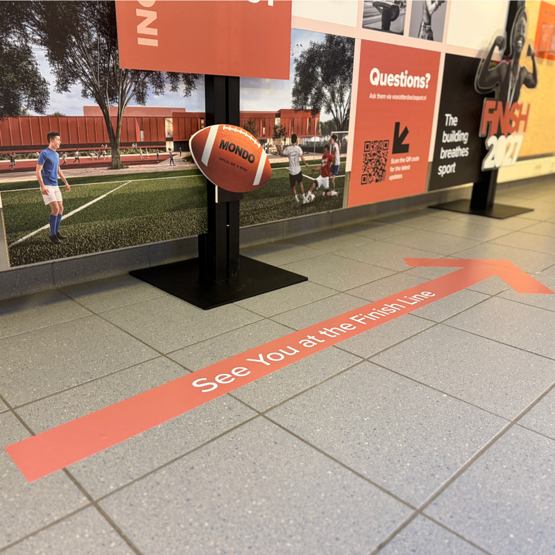 Rode pijl met tekst ‘See You at the Finish Line’ op grijze vloer, richting wand met sportcentrum visuals en QR-code.