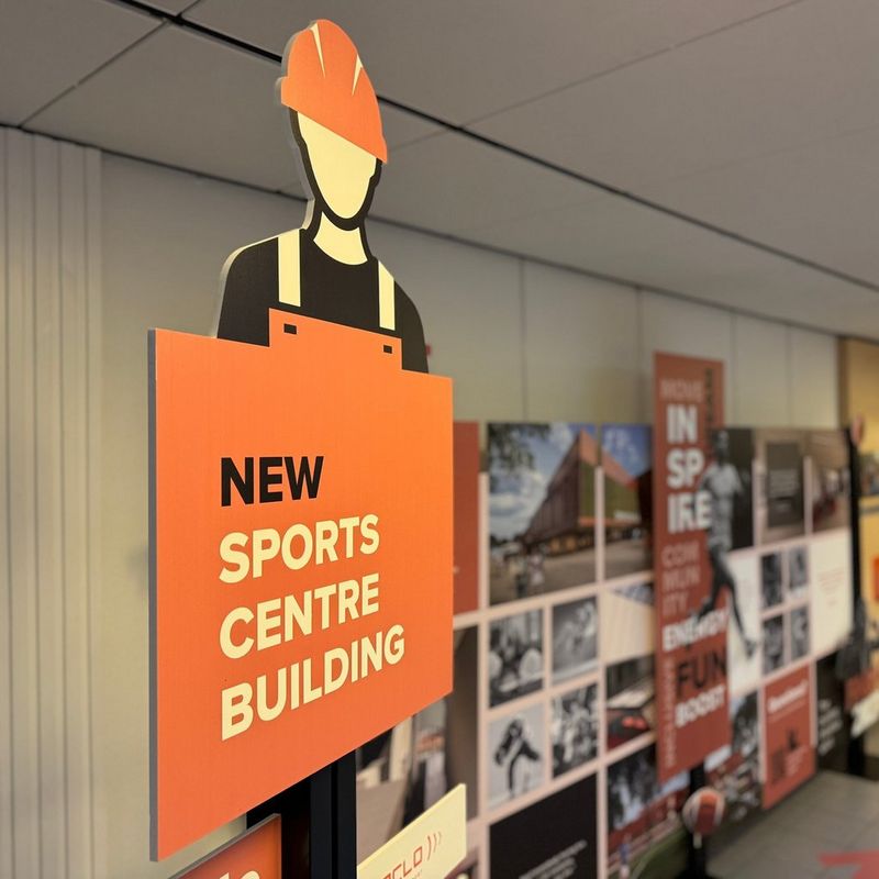 Promotiebord voor nieuw sportcentrum in aanbouw, met illustratie van bouwvakker en tekst ‘New Sports Centre Building’, op de achtergrond inspirerende sportfoto’s en architectuurontwerpen
