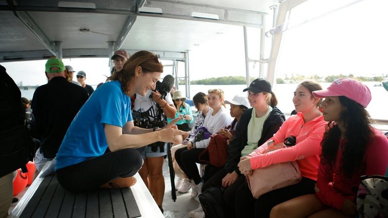 Professor Catherine Macdonald sits cross-legged on the deck of a boat in front of seated students who smile at her as she talks to them.