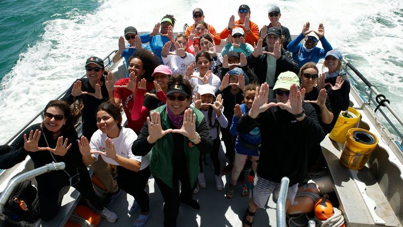 Over twenty crew and youngsters stand at the rear of a boat as it speeds through the water. They are all smiling and holding their hands up, fingers splayed and both thumbs touching.