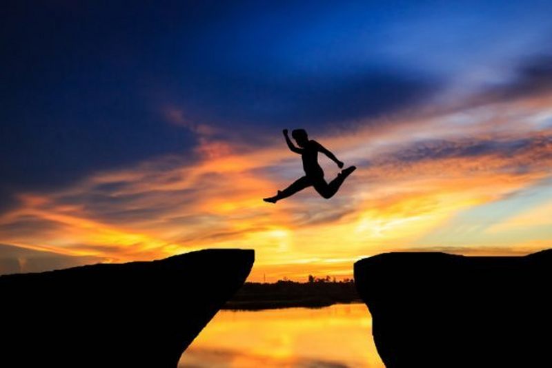 The silhouette of a person leaps between two rocky ledges against a dramatic orange and blue sunset sky reflected on the water below.