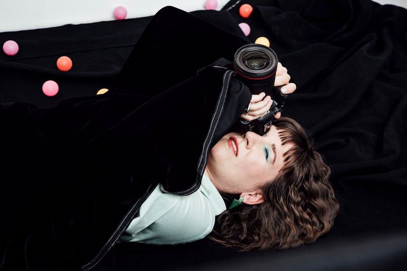 Una donna sdraiata di schiena su un lenzuolo nero e sotto a una coperta dello stesso colore, con una fotocamera vicino al viso.