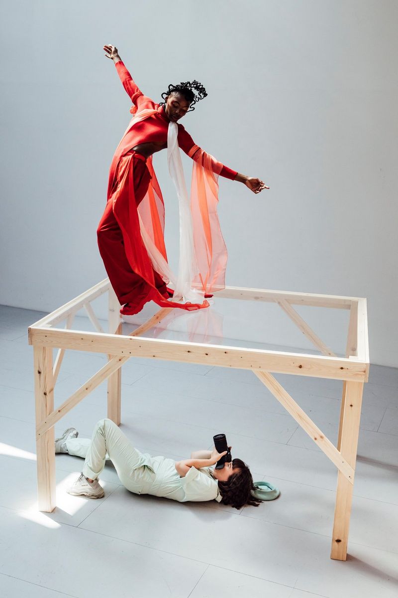 Una ballerina con un morbido abito rosso posa sopra un tavolo in plexiglass chiaro, mentre la fotografa Eliška Sky la immortala dal basso, attraverso il plexiglass.