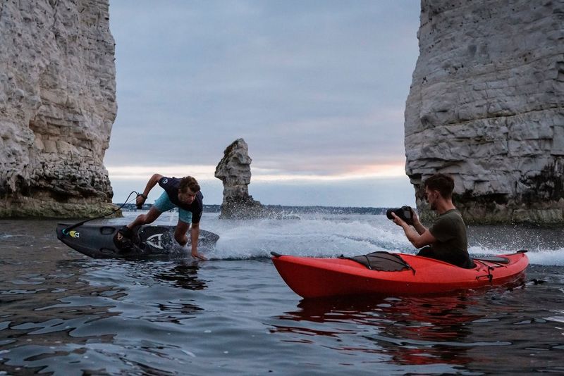 Surfista che si inclina sul lato di un jetboard, in posizione quasi parallela rispetto all'acqua, mentre guarda un fotografo in un kayak rosso che immortala la scena da vicino.
