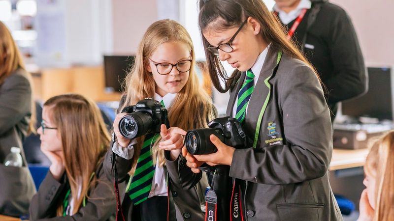 Two girls in a classroom, wearing school uniform and holding Canon cameras.