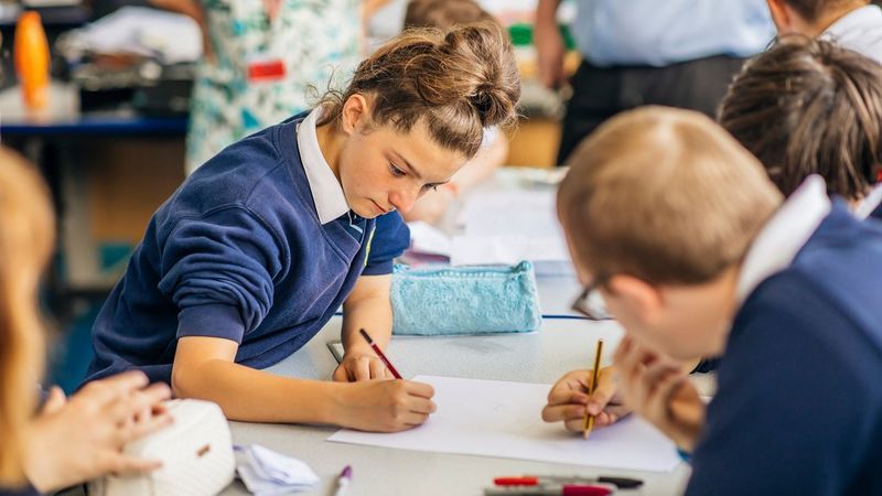 Young people in school uniform sit at a desk drawing on paper.