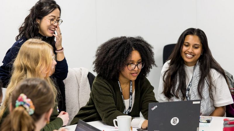 A group of young women smile and laugh while working together at a laptop.