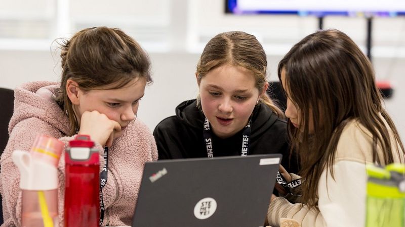 Three young girls huddle closely together as they focus on a laptop screen.