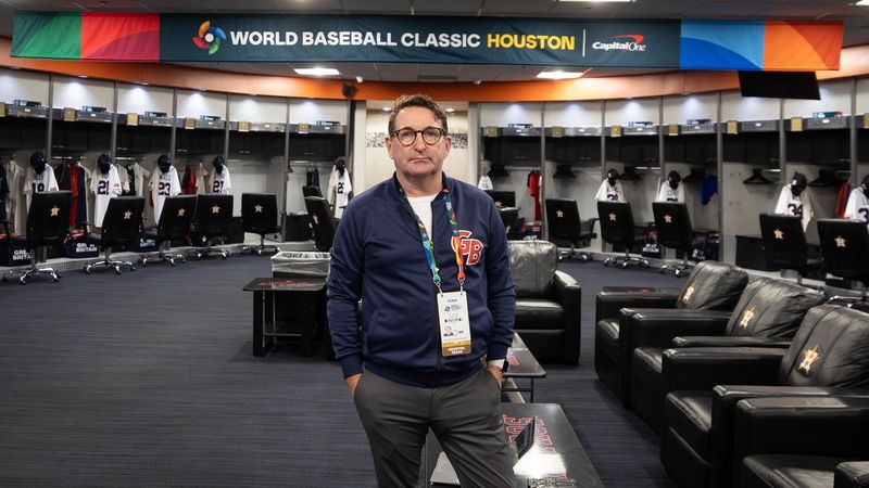 Paul Stodart stands in the centre of a professional baseball locker room, wearing a blue Great Britain baseball jacket and a credential lanyard. A banner above him reads ‘World Baseball Classic Houston’.