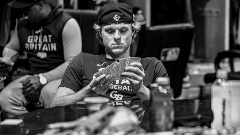 A baseball player in a Great Britain jersey looks intently at a hand of playing cards while sat in a locker room.