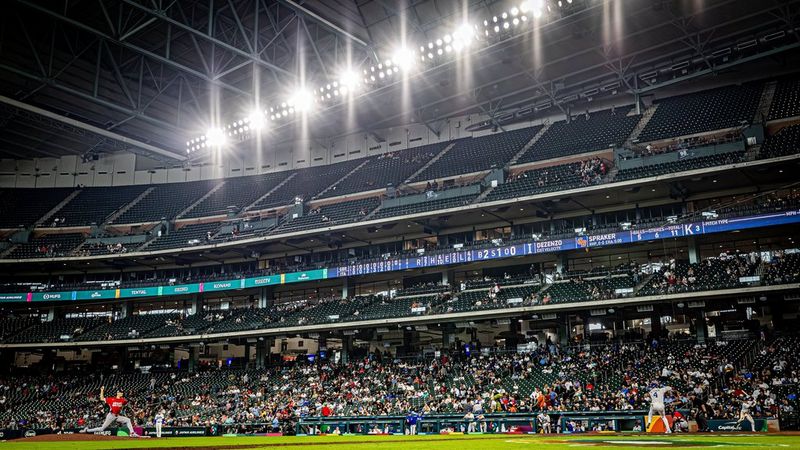 A baseball game at a crowded indoor stadium with bright floodlights and multiple tiers of seating. A pitcher in a red jersey is in mid-delivery on the field.