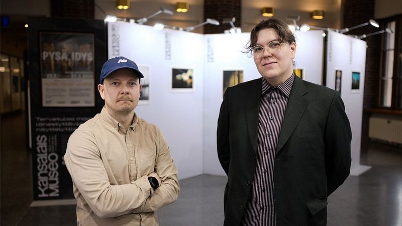 Valokuvaajat Simo Tolvanen ja Pekka Välitalo katsovat kohti kameraa. Valokuvanäyttely näkyy heidän taustallaan.