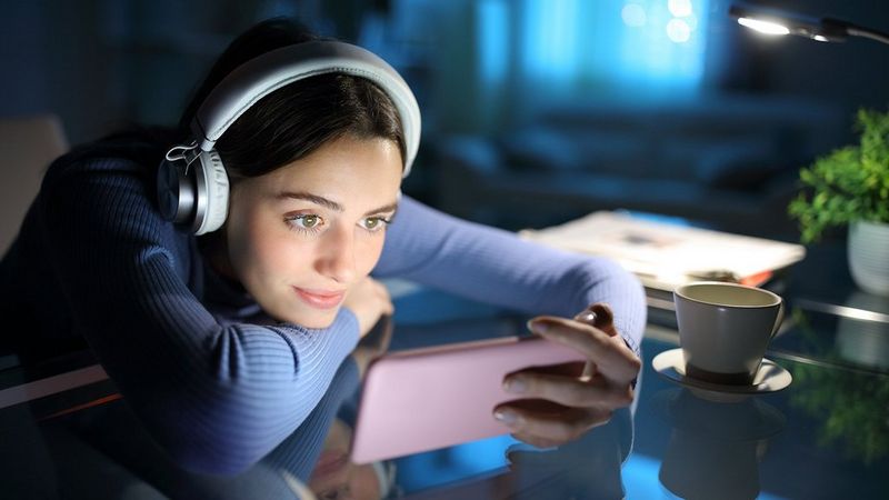 A woman in headphones rests her chin on her arm, which is on a table. In the other hand she holds a phone, landscape, as though watching a video.