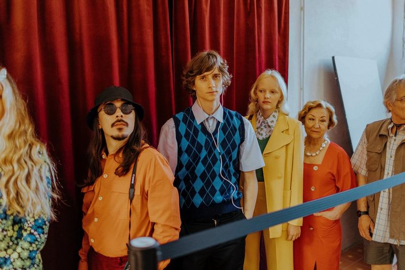 Een rij mensen van verschillende leeftijden en in kleding met een vintage-uitstraling staat voor een gordijn van rood fluweel.