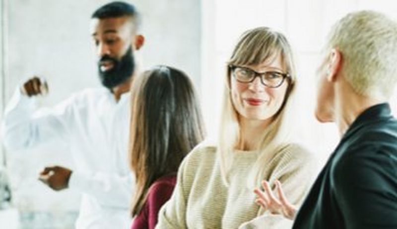 Four office workers stand in pairs chatting, with two gesturing with their hands.