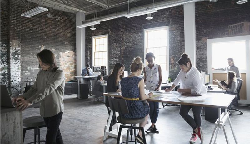 Casual exposed-brick office environment, with one group crowded round a table and others working at laptops and PCs.