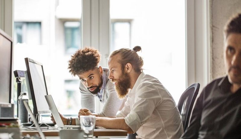 Two bearded men gather round a computer screen, while an out of focus man edges into the frame from the right.