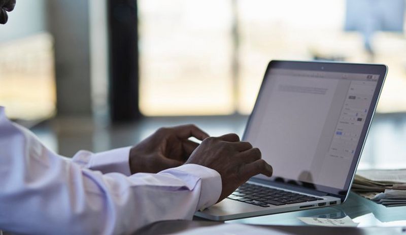 Close-up shot of a man in a crisp white shirt typing into a document on a laptop, with papers piled behind the computer.