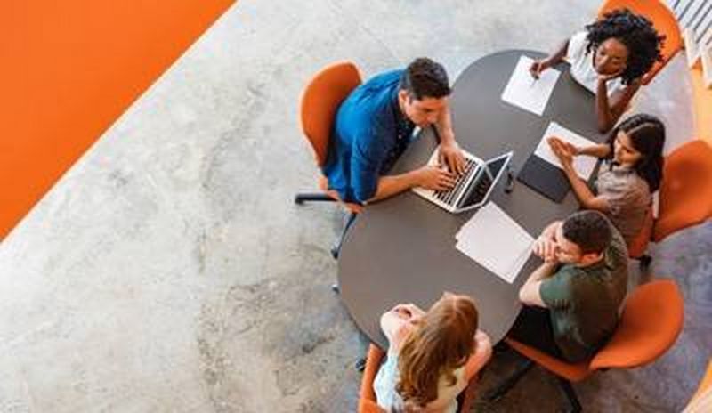 Overhead shot of office workers sat round an oval-shaped table on orange chairs, spread out with papers and a laptop computer.