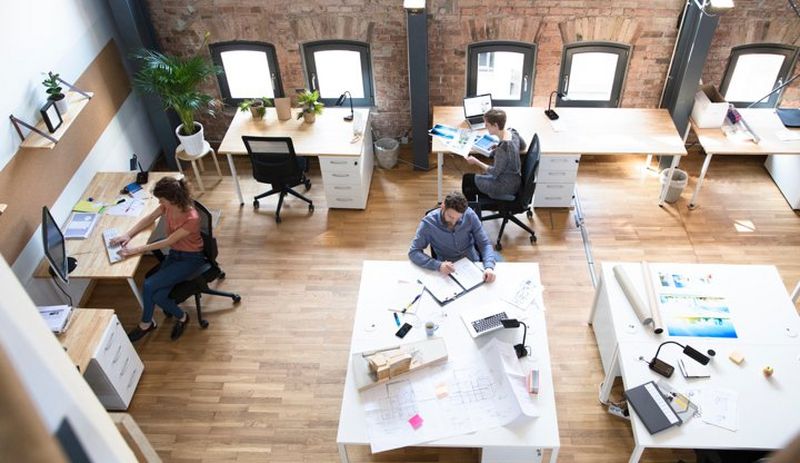 Wide overhead shot of a creative office layout with people working at desks adorned with computers, lamps and brochures.