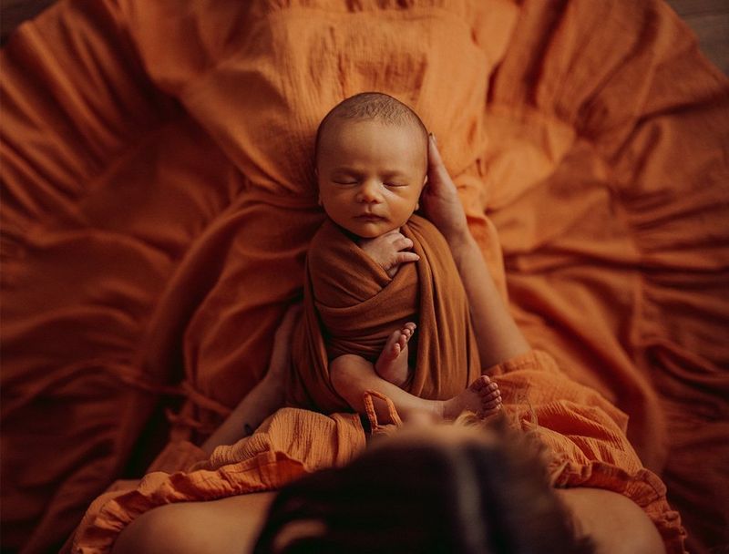 A baby swaddled in an orange fabric held by the mother.