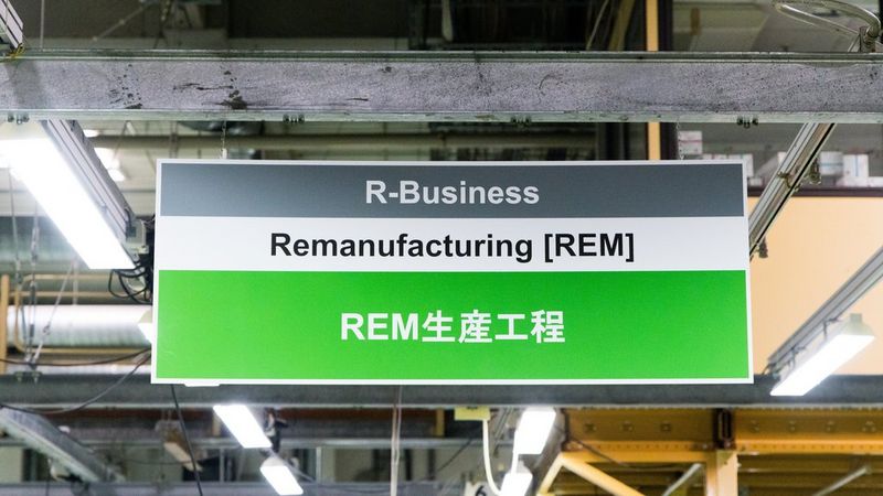 A facility sign hanging from a warehouse ceiling reading ‘R-Business Remanufacturing [REM]’ in English and Japanese.