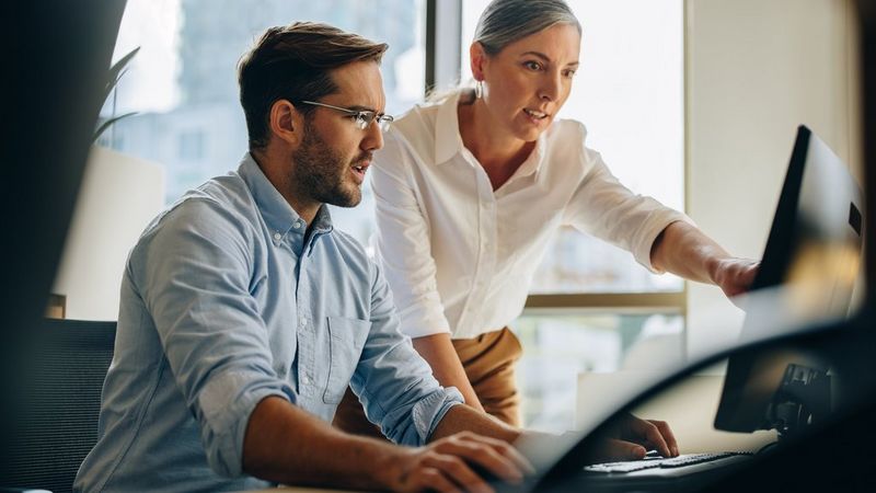 A man and a woman are at a desk looking at a computer screen. The woman points at the screen as she talks to the man.