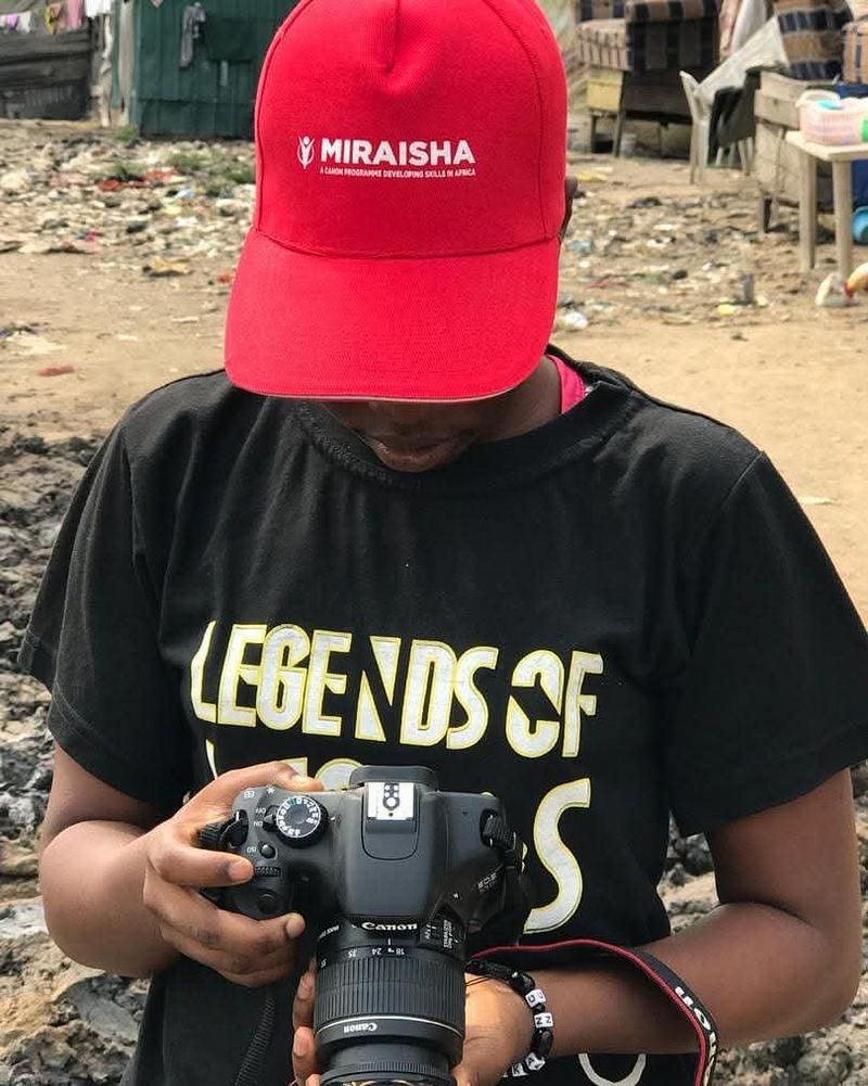 Temitope wears a red baseball cap and a black t-shirt is holding a Canon camera and looking down at the camera's screen. The red cap has a logo that says "MIRAISHA: A FILM PROGRAMME DEVELOPING SKILLS IN AFRICA".