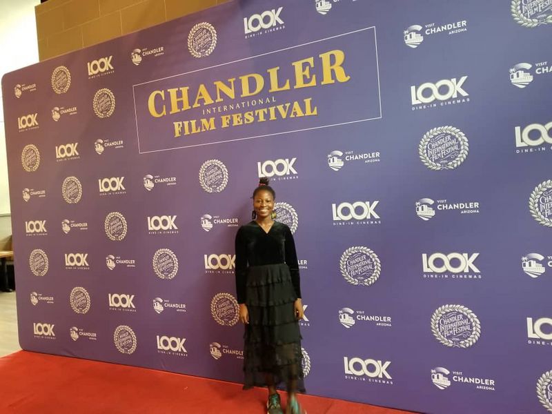 Temitope stands on a red carpet wearing a long black dress and green shoes. The banner behind her bears the logo of the Chandler International Film Festival.  