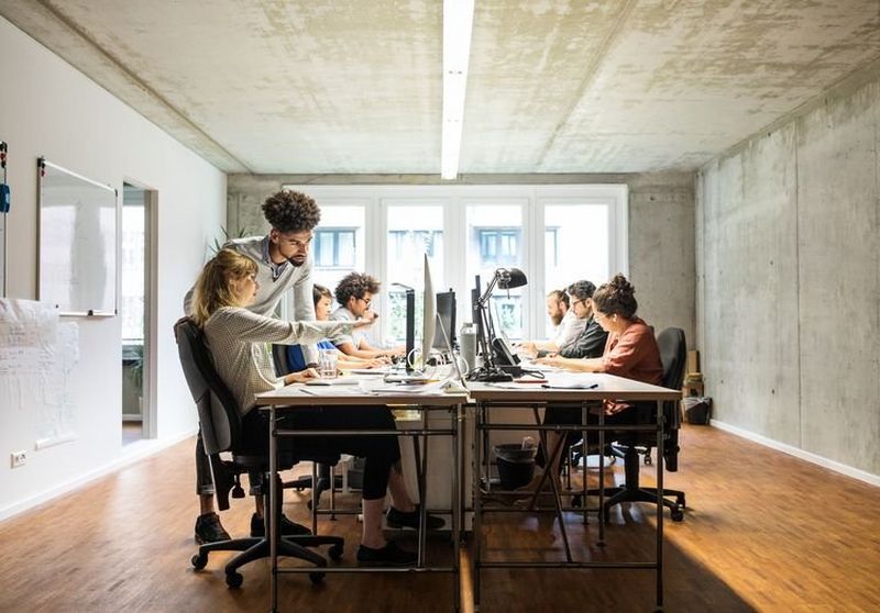 A woman points out something to a colleague standing next her as a team sits round a desk working at their computer screens.