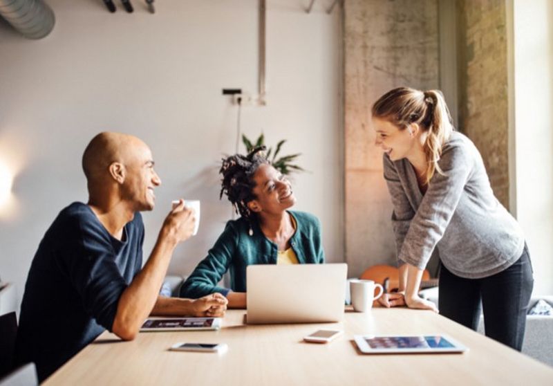 Woman in grey jumper leans hands on table, chatting with a bald man and a woman wearing a green cardigan who sit in front of a laptop, tablet device and phone.