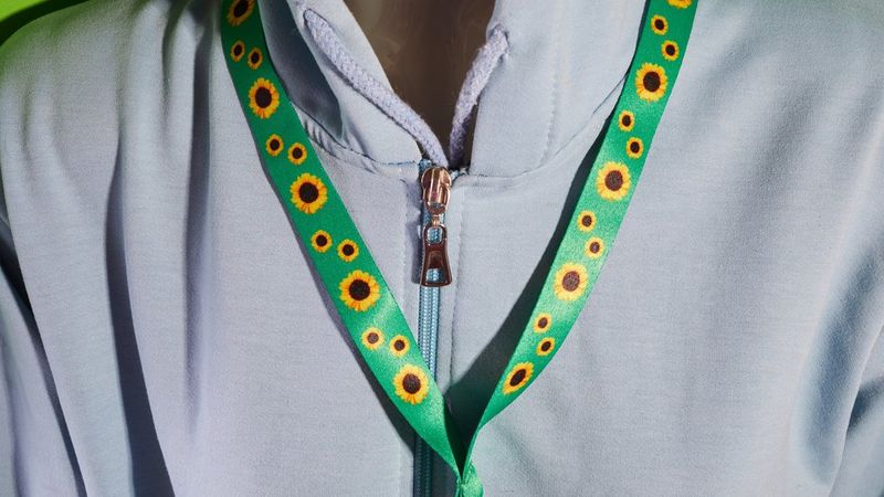 Die Brust einer Person in einer Jacke mit Reißverschluss, die ein grünes, mit Sonnenblumen verziertes Schlüsselband trägt.