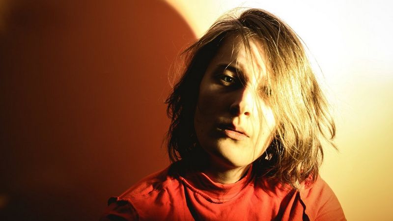 An overexposed photograph of the head and shoulders of a woman. She has shoulder length hair that falls across one eye and is wearing a red top. On the left is her shadow, which looks brown. On the right the wall appears a golden yellow.