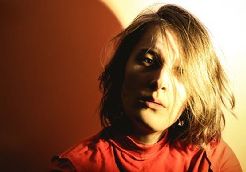 An overexposed photograph of the head and shoulders of a woman. She has shoulder length hair that falls across one eye and is wearing a red top. On the left is her shadow, which looks brown. On the right the wall appears a golden yellow.