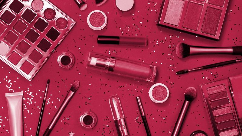 A selection of cosmetics and make-up brushes lying on a crimson/magenta table. There are also sparkles and glitter on the table.