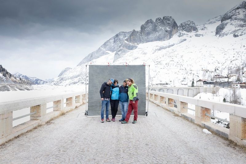 Four people stand in front of a grey photographer’s backdrop with their heads bowed and arms around each other. Beyond the backdrop are huge snow-covered mountains. 