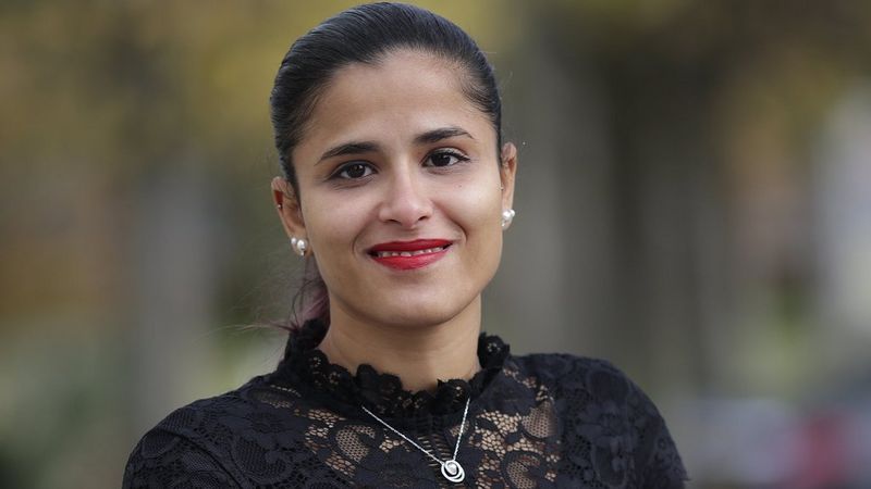 A head and shoulders image of a smiling dark haired, dark skinned woman. Her hair is tied back and she wears pearl earrings. Over her black lace top is a silver necklace.