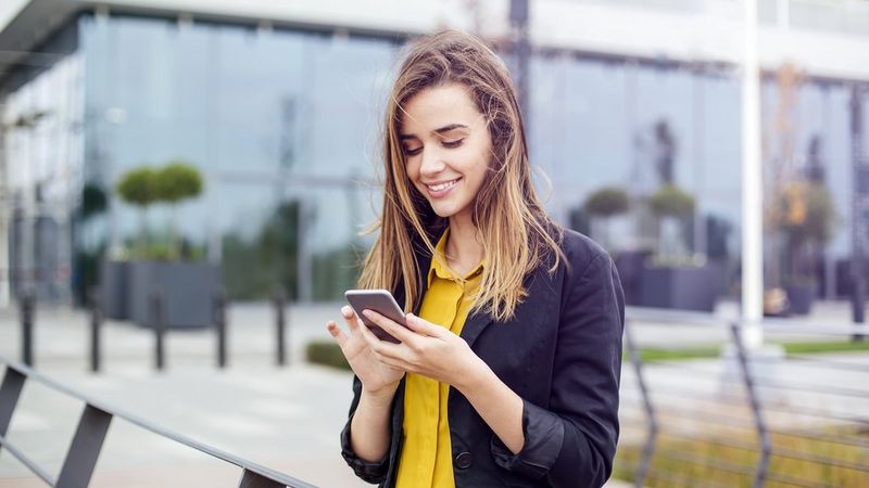 A young white woman with long hair, wearing a yellow shirt and blue blazer stands outside an office and smiles as she checks her phone.