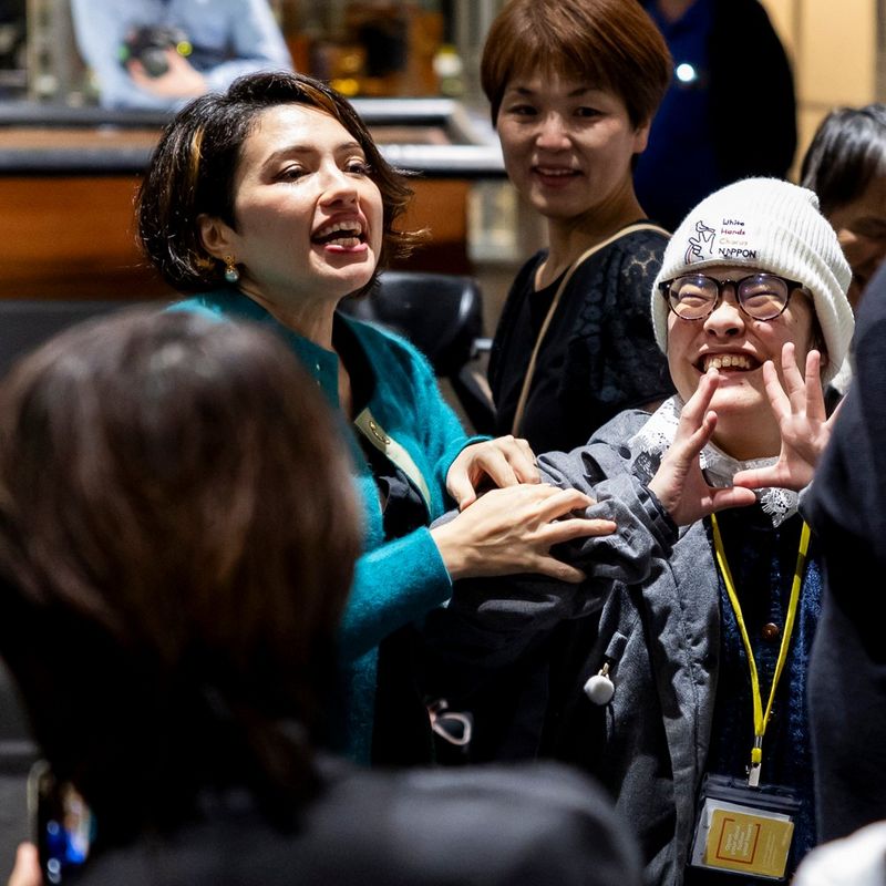 A dark haired woman, who appears to be mid-sentence, stands among a group of people. Next to her is a young person in a white hat and glasses who holds their hands up as though using sign language and is smiling widely.