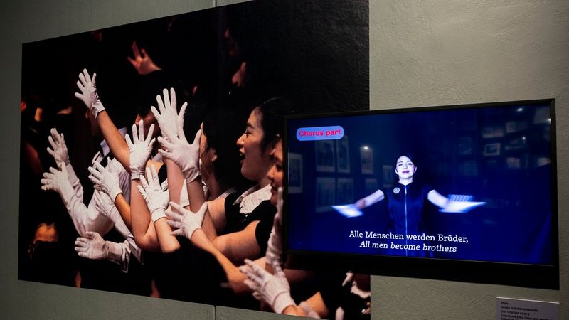 A printed photograph of many people, facing left, with their white-gloved hands raised. Beside it is a video screen showing a woman with her arms outstretched. The caption below her reads: Alle Menschen warden Brüder, All men become brothers.